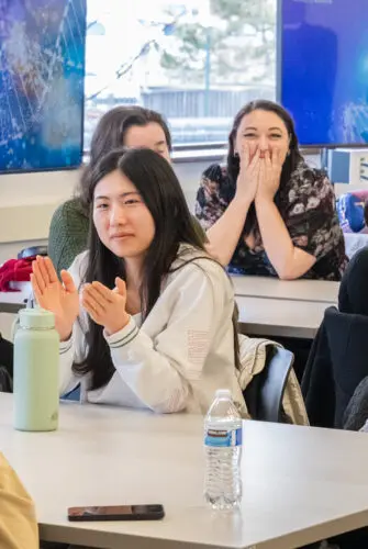 A group of approximately 10 students sit at tables in a classroom. Several are clapping, and two are covering their mouths in surprise.
