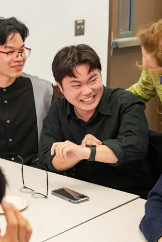 Two men wearing black shirts sit at a table talking to a standing woman in a green jacket. The man on the left is wearing glasses and smiling, and the man on the right is grinning and pinching his own arm.
