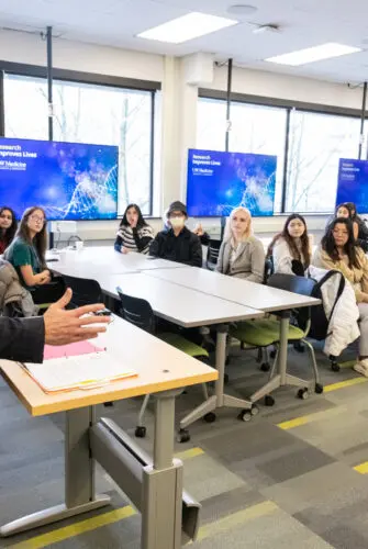 A man wearing glasses and a suit gestures with his right hand while addressing a classroom full of students. A cameraman is visible behind him.