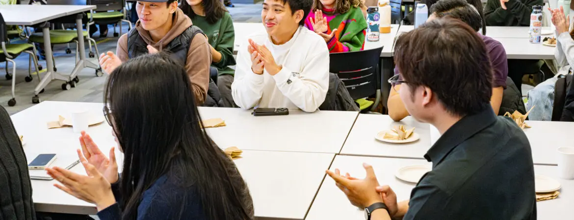 A classroom full of students sits at tables and applauds.