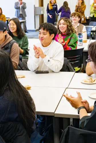 A classroom full of students sits at tables and applauds.