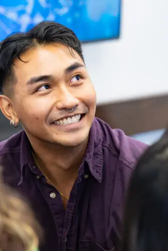 A man with dark hair wearing a purple shirt sits at a table, smiling, with his face looking to the right.
