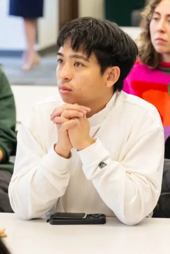 A man in a white long-sleeved shirt sits at a table, with other students visible next to and behind him. His hands are clasped in front of him and he is looking to the left.