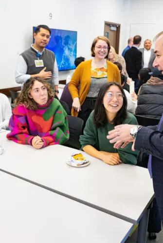 A man in a dark suit and a blue tie stands at the right, talking to a group of students sitting at a table. He is gesturing as he speaks.