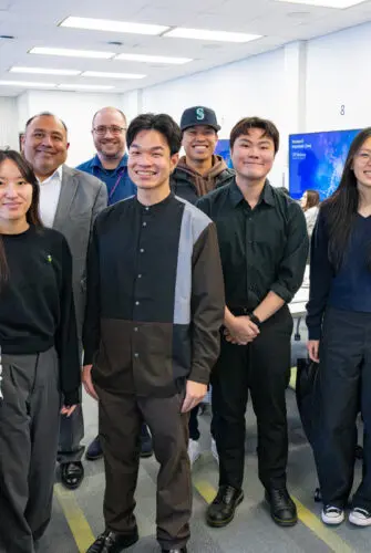 A group of 10 people of varying ages, most wearing dark clothing but one wearing a white shirt, stand facing the camera and smiling.