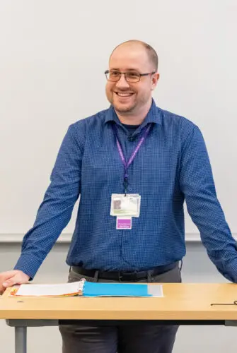 A man in a blue checked shirt stands at a lectern in front of a whiteboard. He is resting his hands on the lectern and smiling.
