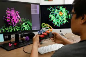 A researchers at the UW Medicine Institute for Protein Design, holding a 3-D model of a protein and looking at two monitors with computer-generated protein images