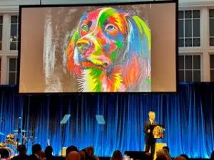 A colorful painting of a dog, by Jessie Owen, shown on a large projector. A man speaks behind a podium below the screen.
