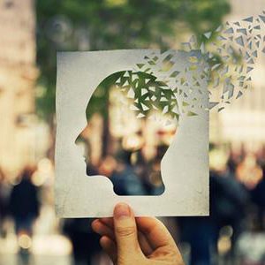 A hand holding a piece of paper with a cut out of person's face in silhouette, with smaller pieces of paper fragmenting off from the person's brain. Behind the piece of paper is a blurry image of a busy street.
