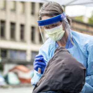 A healthcare provider in personal protective equipment administering a COVID-19 nasal swab to a patient outside.